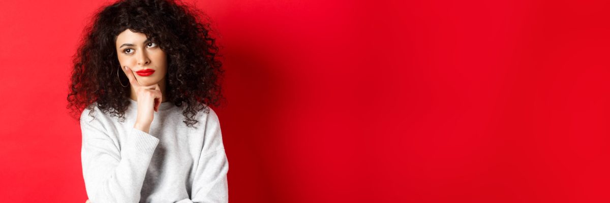 Grumpy young woman with curly hair, looking annoyed or bored at empty space, standing pensive and sad on red background.