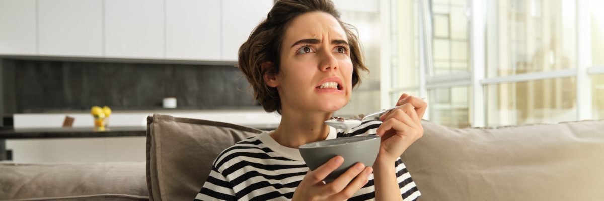 Image of woman eating breakfast in front of tv, looking at screen with worried face, holding bowl of cereals with milk and a spoon, spending time in living room.